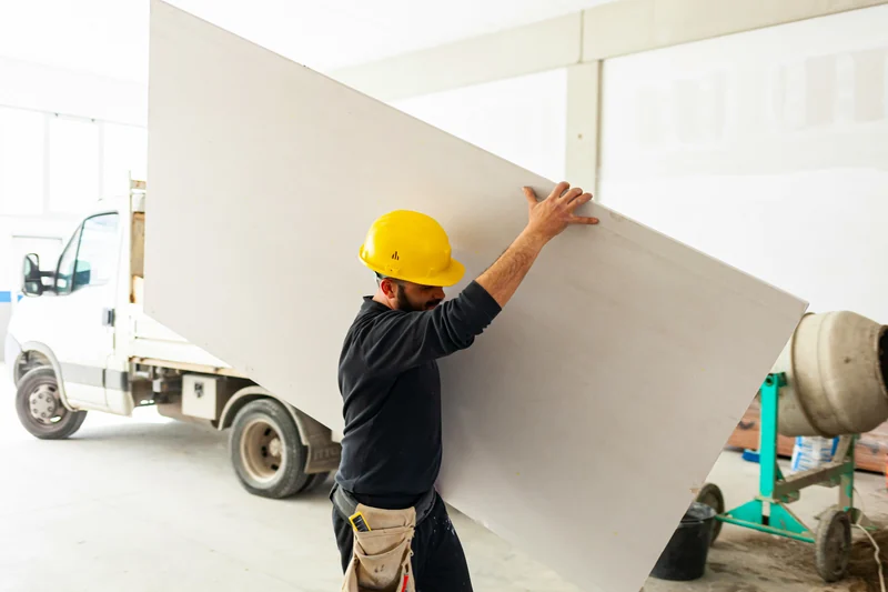 Worker carrying a drywall sheet in from a delivery truck