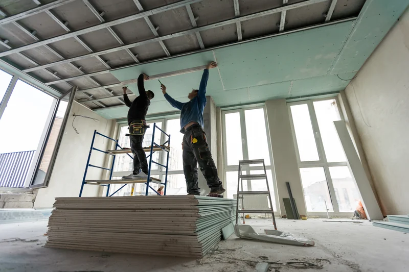 Workers on scaffolding beside a stack of board in a tall commercial room
