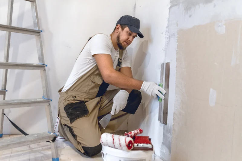 Worker kneeling to patch a low wall beside a paint roller and tray
